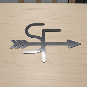 Op de afbeelding: Metalen pijl-vormige wanddecoratie met de letters "SF" in het ontwerp verwerkt. De pijl wijst naar rechts en het metaal heeft een geborstelde afwerking. Op een lichte houten achtergrond.