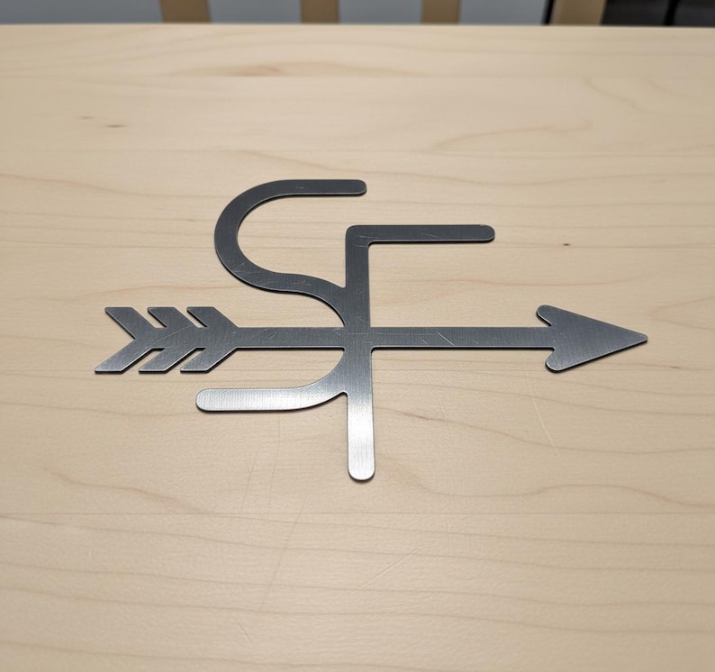 Op de afbeelding: Metalen pijl-vormige wanddecoratie met de letters "SF" in het ontwerp verwerkt. De pijl wijst naar rechts en het metaal heeft een geborstelde afwerking. Op een lichte houten achtergrond.