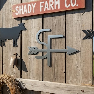 Op de afbeelding: Houten wanddecoratie met een metalen koe-silhouet, een bel en een metalen pijl met de letters "SF". Een rood bord bovenaan leest "SHADY FARM CO.". De decoratie is op een houten muur gemonteerd.