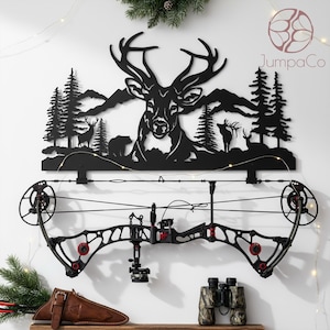 Peut inclure: Décoration murale en métal noir représentant une tête de cerf, des montagnes et des arbres, avec un arc à poulies monté en dessous. L'arc a des détails noirs et rouges. Un étui à arc en cuir marron et des jumelles de camouflage sont sur une étagère en bois.