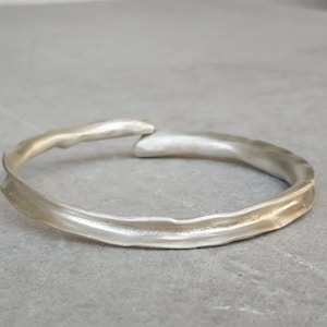 May include: A silver-colored, open-ended bracelet with an organic, textured design. The bracelet has a smooth, polished finish and a slightly irregular shape. The overall aesthetic is minimalist and modern.