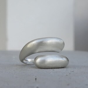 Könnte beinhalten: Ein silberfarbener Ring mit einem einzigartigen, organischen Design. Der Ring hat zwei abgerundete, glatte und längliche Formen, die sich um den Finger zu wickeln scheinen. Das Metall hat eine matte Oberfläche, die ihm ein weiches, diffuses Aussehen verleiht. Der Ring ist vor einem neutralen Hintergrund platziert.