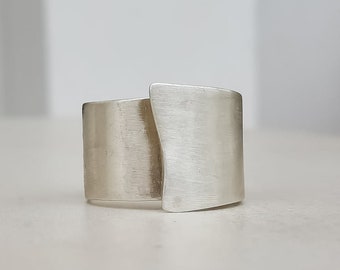 May include: A silver ring with a modern design. The ring features two rectangular bands that overlap, creating a unique and contemporary look. The metal has a brushed finish, giving it a matte appearance.