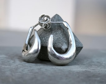 May include: A pair of silver-toned, teardrop-shaped earrings. The earrings have a smooth, polished finish and a post-and-clutch closure. They are displayed on a gray stone surface, showcasing their reflective quality. The earrings are a simple, elegant jewelry item.