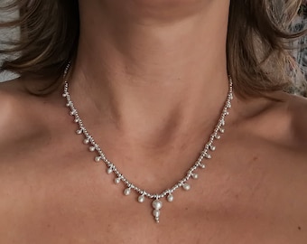 May include: A silver necklace with a delicate design. The necklace features small, round beads and teardrop-shaped pendants. The necklace is worn around the neck and is a piece of jewelry.