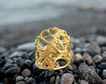 May include: A gold-colored ring with an open, web-like design. The ring is set against a backdrop of small, smooth, dark gray and brown stones, with a blurred ocean in the background. The ring's intricate pattern creates a unique, textured appearance.