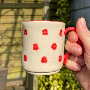 May include: A cream-coloured ceramic mug with a red handle and interior, adorned with small red floral patterns. The mug is held in a hand, with a blurred background of foliage. The mug is suitable for hot beverages.