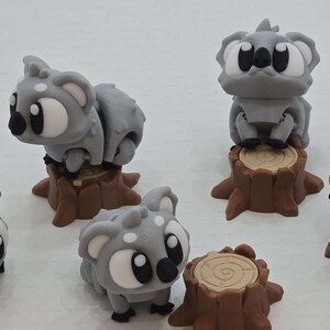 May include: Six gray koala figurines with black eyes and brown tree stump bases. The koalas are in various poses, some sitting on the stumps and others standing.
