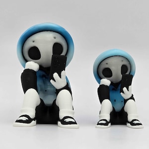 May include: Two figurines of a skeleton-like character wearing a blue hoodie, black pants, and white shoes, each holding a black smartphone. The figures are sitting with their legs crossed, with one figure slightly smaller than the other.