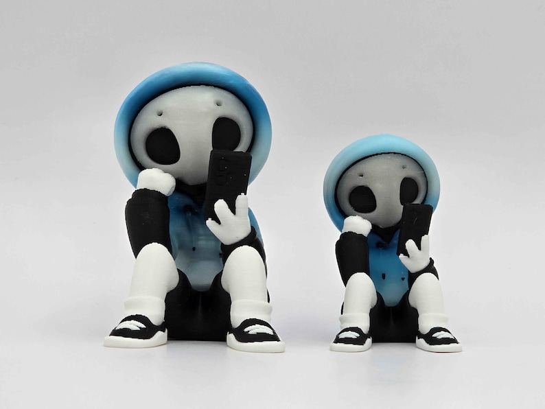 May include: Two figurines of a skeleton-like character wearing a blue hoodie, black pants, and white shoes, each holding a black smartphone. The figures are sitting with their legs crossed, with one figure slightly smaller than the other.