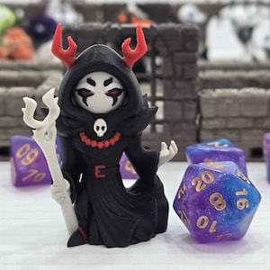 May include: A black figurine of a hooded character with red horns, holding a white staff. The figure has a skull on its chest and is surrounded by colorful dice, suggesting a tabletop gaming theme.