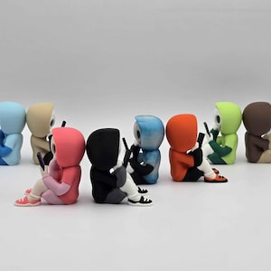 May include: A collection of colorful figurines, each depicting a hooded figure seated and holding a smartphone. The figures are in various colors, including blue, tan, pink, black, blue, orange, green, and brown. They are wearing sneakers.