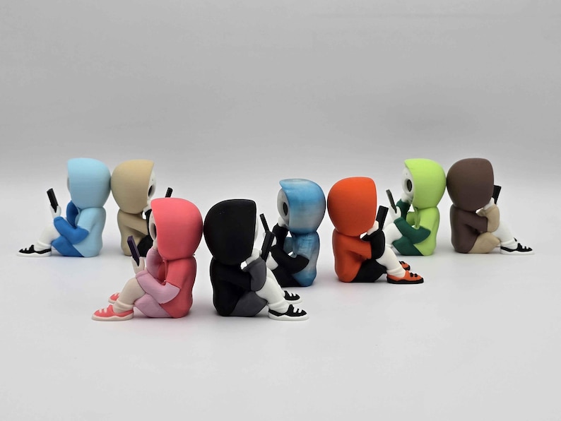 May include: A collection of colorful figurines, each depicting a hooded figure seated and holding a smartphone. The figures are in various colors, including blue, tan, pink, black, blue, orange, green, and brown. They are wearing sneakers.
