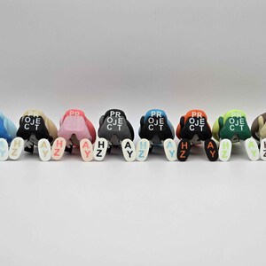 May include: A collection of colorful figurines, each with a unique color scheme and the word "PROJECT" printed on the back. Each figurine has the letters "HAY" and "AYZ" printed on the front. The figurines are arranged in a row.