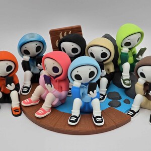 May include: A group of stylized figurines, each wearing a different colored hoodie and holding a smartphone. The figures are seated on a circular platform with a blue and brown design, suggesting a social gathering or digital interaction theme.
