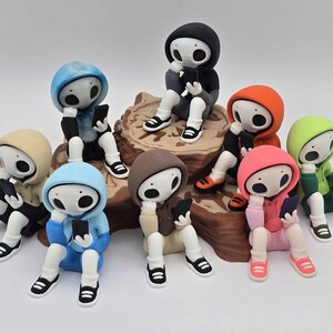 May include: A collection of whimsical figurines, each in a different colored hoodie, sitting and holding a smartphone. The figurines have white bodies, black shoes, and large, expressive eyes. They are arranged on a brown, textured base.