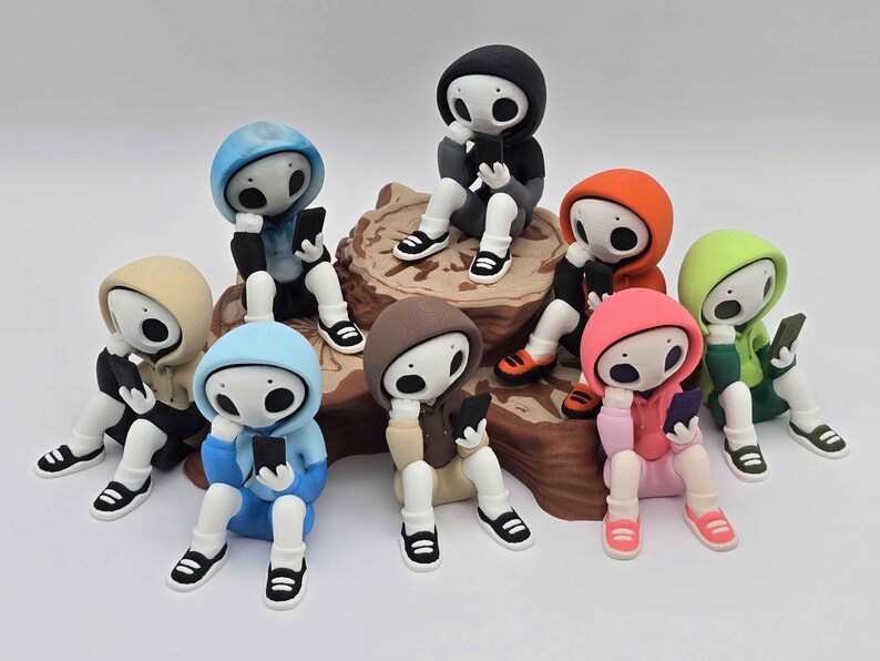 May include: A collection of whimsical figurines, each in a different colored hoodie, sitting and holding a smartphone. The figurines have white bodies, black shoes, and large, expressive eyes. They are arranged on a brown, textured base.