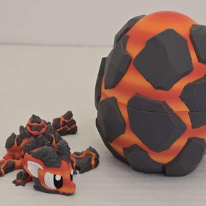 May include: A 3D-printed dragon and egg set. The dragon is orange, black, and gray, with a stylized design. The egg is a sphere with a lava-like pattern of orange and black. The set is designed to look like a volcanic theme.