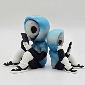 May include: Two figurines of hooded figures in blue and black, each holding a black smartphone. The figures are seated with white legs and black and white sneakers. The figures have a skull-like face.