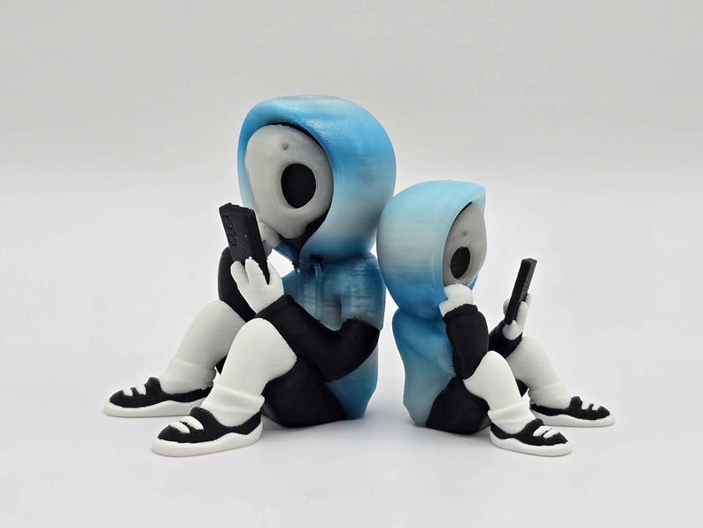 May include: Two figurines of hooded figures in blue and black, each holding a black smartphone. The figures are seated with white legs and black and white sneakers. The figures have a skull-like face.