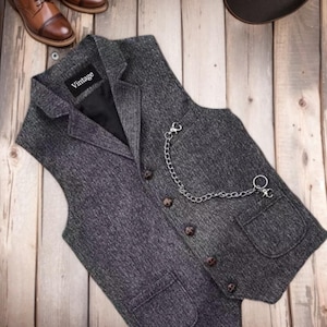 May include: A charcoal grey herringbone waistcoat with a notched lapel, five dark buttons, and two front pockets. A silver-toned metal chain is attached to the chest area. A black label with the word "Vintage" is sewn inside the collar area.