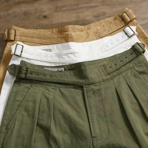 Puede incluir: Tres pantalones en verde oliva, blanco y beige, cada uno con dise&ntilde;o de cintura alta, correa de ajuste lateral y hebilla de metal. Los pantalones presentan una construcci&oacute;n frontal con pliegues y una etiqueta de marca visible.