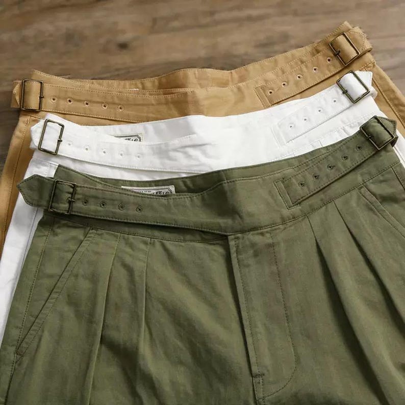 Puede incluir: Tres pantalones en verde oliva, blanco y beige, cada uno con dise&ntilde;o de cintura alta, correa de ajuste lateral y hebilla de metal. Los pantalones presentan una construcci&oacute;n frontal con pliegues y una etiqueta de marca visible.