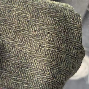 May include: A piece of woven fabric featuring a dark olive green and black herringbone pattern. The material has a visible textured surface with a dense, fibrous weave. A small section of a red and green striped border is visible at the bottom edge.