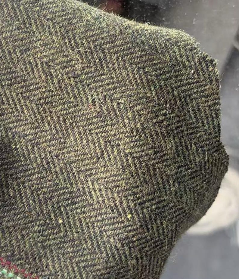 May include: A piece of woven fabric featuring a dark olive green and black herringbone pattern. The material has a visible textured surface with a dense, fibrous weave. A small section of a red and green striped border is visible at the bottom edge.