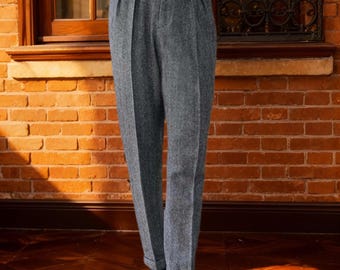 May include: Gray herringbone tweed trousers with a high waist and pleated front. The pants have a tailored fit and are cuffed at the ankles. The background features a brick wall and a wooden floor.