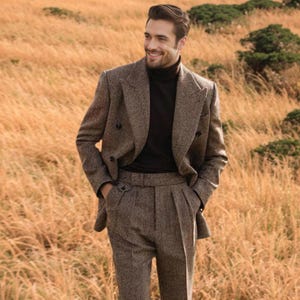 May include: A brown herringbone suit with a double-breasted jacket and pleated trousers. The suit is worn over a black turtleneck. The background features tall, dry grass and a natural setting.