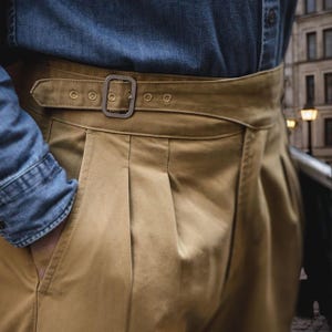 May include: Khaki-colored pants with a matching belt and a silver buckle. The pants have pleats and a high waist. A denim shirt is partially visible in the image.