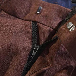 Pu&ograve; includere: Primo piano della cintura e della cerniera di pantaloni marroni. I pantaloni hanno una cerniera nera e una chiusura in metallo. Il tessuto sembra essere una trama strutturata. L'immagine mostra i dettagli della costruzione dell'indumento.