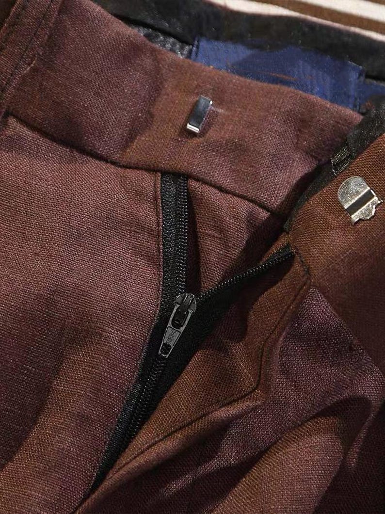 Pu&ograve; includere: Primo piano della cintura e della cerniera di pantaloni marroni. I pantaloni hanno una cerniera nera e una chiusura in metallo. Il tessuto sembra essere una trama strutturata. L'immagine mostra i dettagli della costruzione dell'indumento.