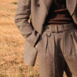 May include: A brown herringbone tweed suit, comprising a blazer and high-waisted trousers. The blazer has a two-button fastening, and the trousers feature a belt. The suit is crafted from a textured fabric, presenting a classic, tailored aesthetic.