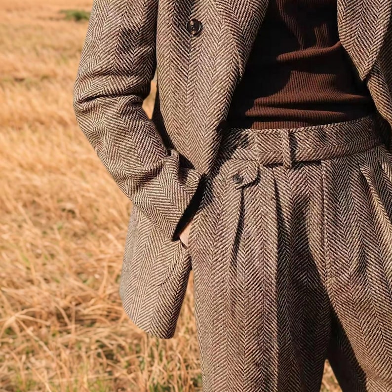 May include: A brown herringbone tweed suit, comprising a blazer and high-waisted trousers. The blazer has a two-button fastening, and the trousers feature a belt. The suit is crafted from a textured fabric, presenting a classic, tailored aesthetic.