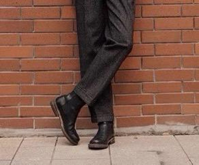 May include: A pair of dark charcoal gray trousers with a subtle textured pattern paired with black leather ankle boots. The trousers feature a straight-leg cut that falls to the ankle, while the boots have a smooth finish and a low heel.