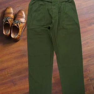 Puede incluir: Pantalones de color verde oliva con dise&ntilde;o de cintura alta, ajustadores de hebilla laterales en la pretina y un par de zapatos oxford de cuero marr&oacute;n con cordones, presentados juntos como un conjunto coordinado de ropa y calzado.