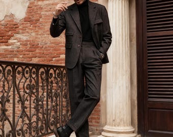 Puede incluir: Un traje gris carbón oscuro compuesto por una americana con bolsillos de solapa y pantalones a juego, usado con un suéter de cuello alto negro, un sombrero fedora marrón y botas de cuero negro. El conjunto presenta una tela texturizada.