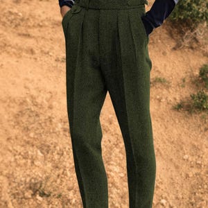 May include: Dark green herringbone trousers with a high waist and pleated front. The trousers have a tailored fit, cuffed hems, and a buttoned waistband. The model is wearing a dark blue long-sleeved shirt.