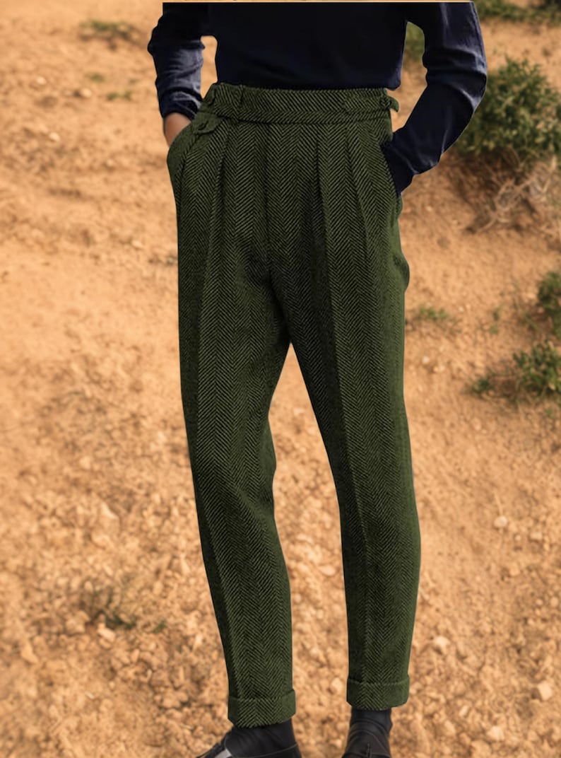 May include: Dark green herringbone trousers with a high waist and pleated front. The trousers have a tailored fit, cuffed hems, and a buttoned waistband. The model is wearing a dark blue long-sleeved shirt.