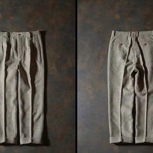 May include: A pair of light brown pleated trousers, displayed front and back. The trousers have a textured fabric, a button closure, and pockets. The back view shows two red buttons. The pants are laid out on a dark brown surface.