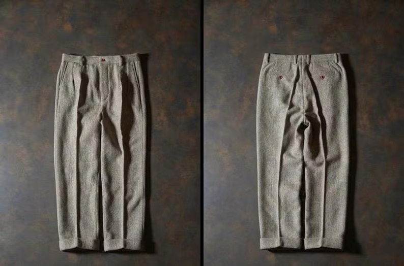 May include: A pair of light brown pleated trousers, displayed front and back. The trousers have a textured fabric, a button closure, and pockets. The back view shows two red buttons. The pants are laid out on a dark brown surface.