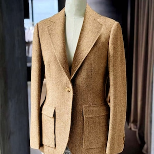 May include: A tan herringbone blazer with two front pockets and a single-button closure. The tailored jacket is displayed on a mannequin, showcasing its classic design and structured fit. The fabric has a subtle pattern.