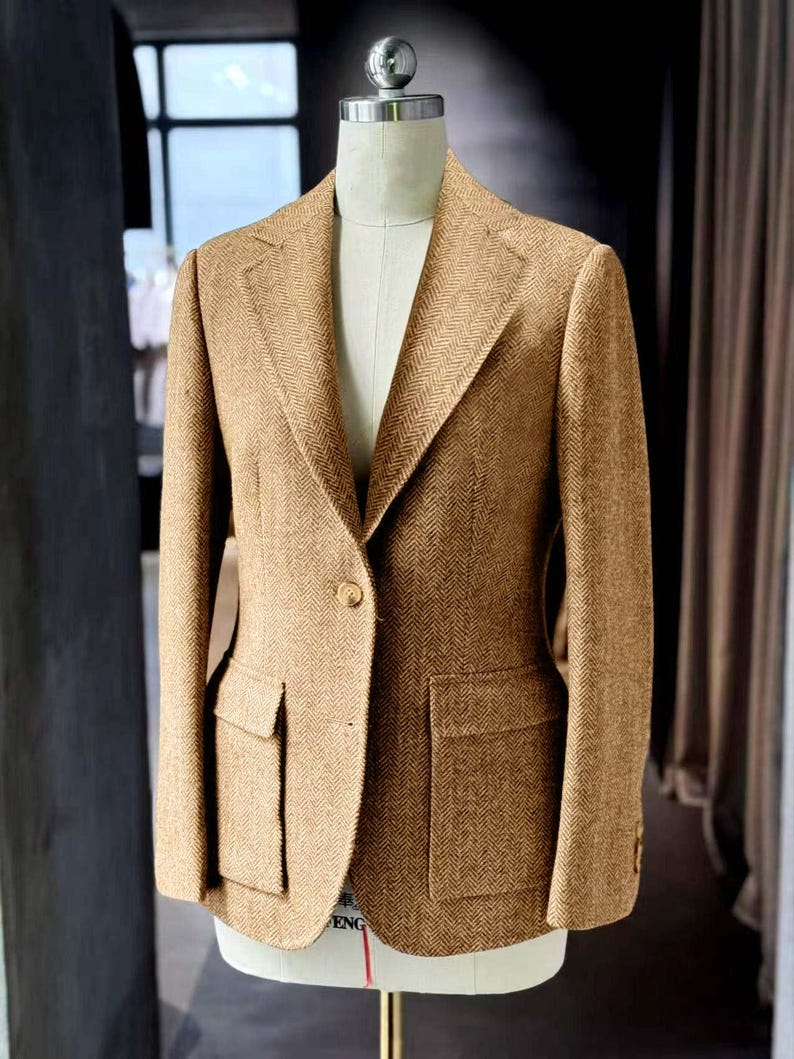 May include: A tan herringbone blazer with two front pockets and a single-button closure. The tailored jacket is displayed on a mannequin, showcasing its classic design and structured fit. The fabric has a subtle pattern.