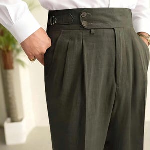 May include: High-waisted olive green trousers featuring a wide waistband with two buttons, a side adjuster strap with a metal buckle, and front pleats. The trousers are paired with a crisp white long-sleeved button-down shirt tucked into the waist.