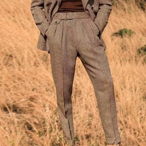May include: A brown tweed suit, comprising a blazer and tailored trousers. The trousers feature a pleated front and a belt. The suit is made from a textured fabric, suitable for formal or business wear.
