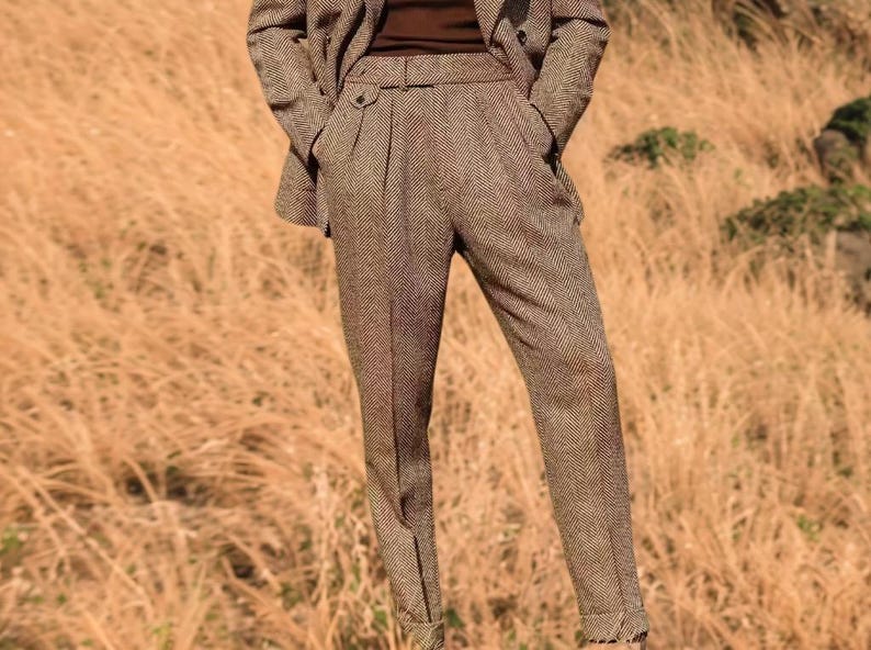 May include: A brown tweed suit, comprising a blazer and tailored trousers. The trousers feature a pleated front and a belt. The suit is made from a textured fabric, suitable for formal or business wear.