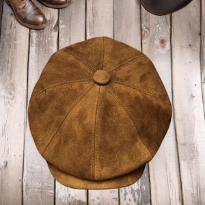 Peut inclure: Casquette de style gavroche en daim marron avec un bouton sur le dessus. La casquette est d'une teinte chaude et terreuse et a une forme arrondie. L'image montre également des bottes en cuir marron et un chapeau marron.