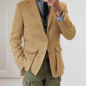 May include: A tan herringbone blazer with two front pockets and a single-button closure. The blazer is paired with a denim shirt, a patterned vest, a dark tie, and olive green cargo pants. The outfit is a blend of textures and colors.